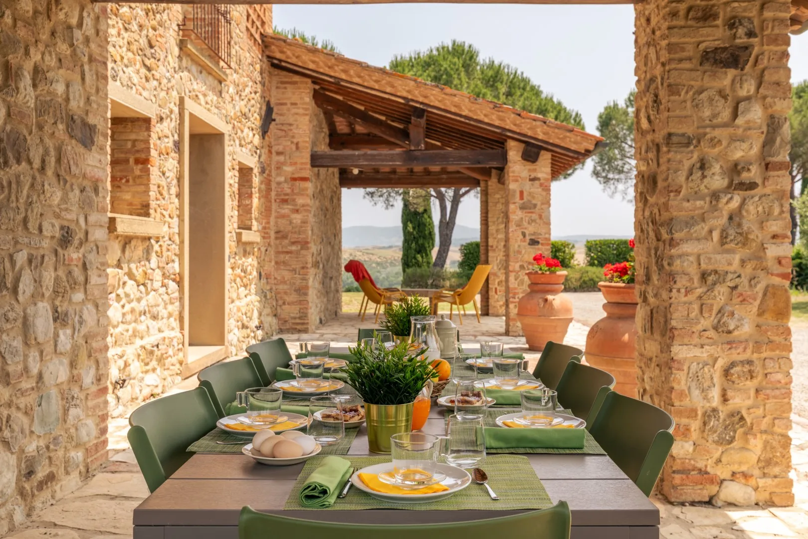 An outdoor dining table is set beneath a stone pergola, with greenery nearby.
