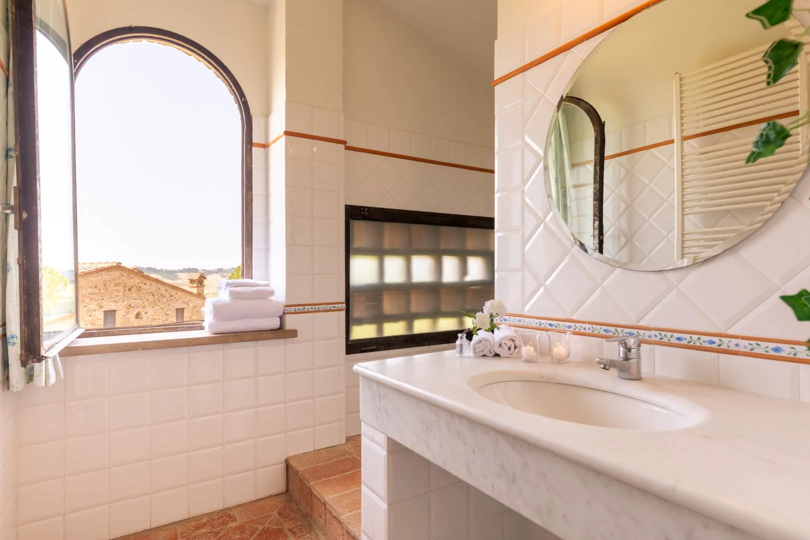 A tiled bathroom with a sink, round mirror, and arched window.