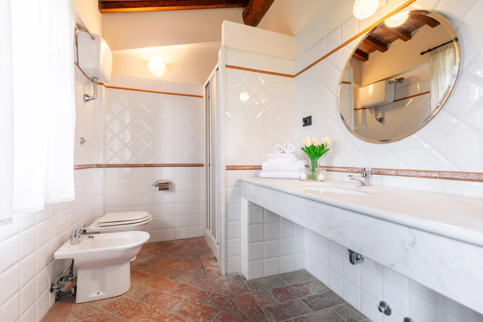 A tiled bathroom with a sink, toilet, and large round mirror.