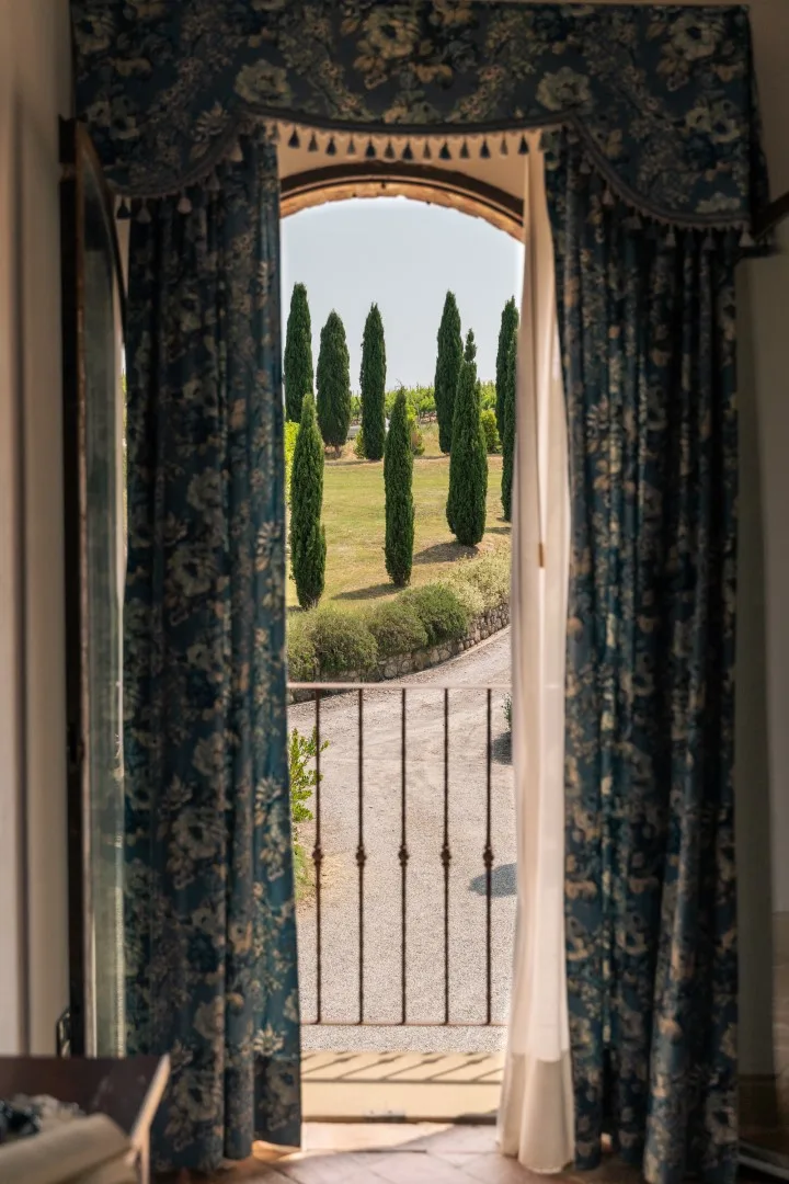 A curtained window opens onto a garden with tall trees and a railing.