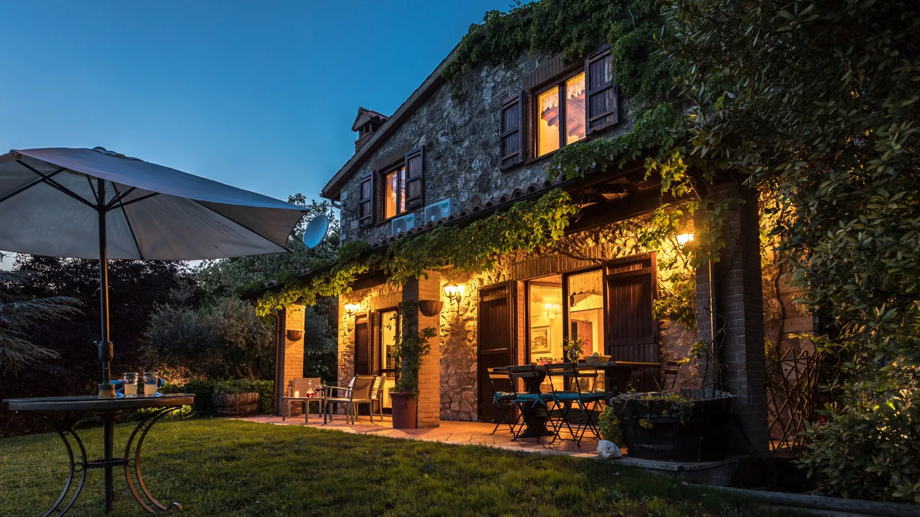 Una casa de piedra con un patio iluminado y asientos al aire libre al anochecer.