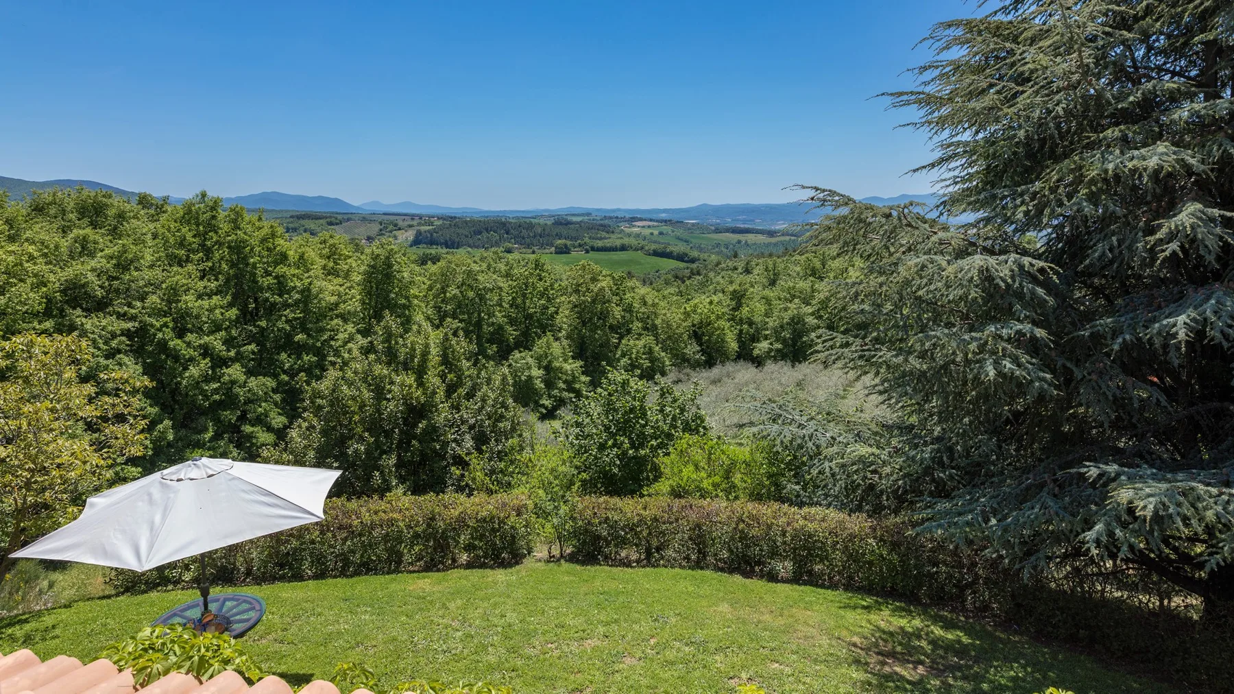 Un jardín de césped tiene vistas a un valle boscoso y colinas lejanas bajo un cielo despejado.