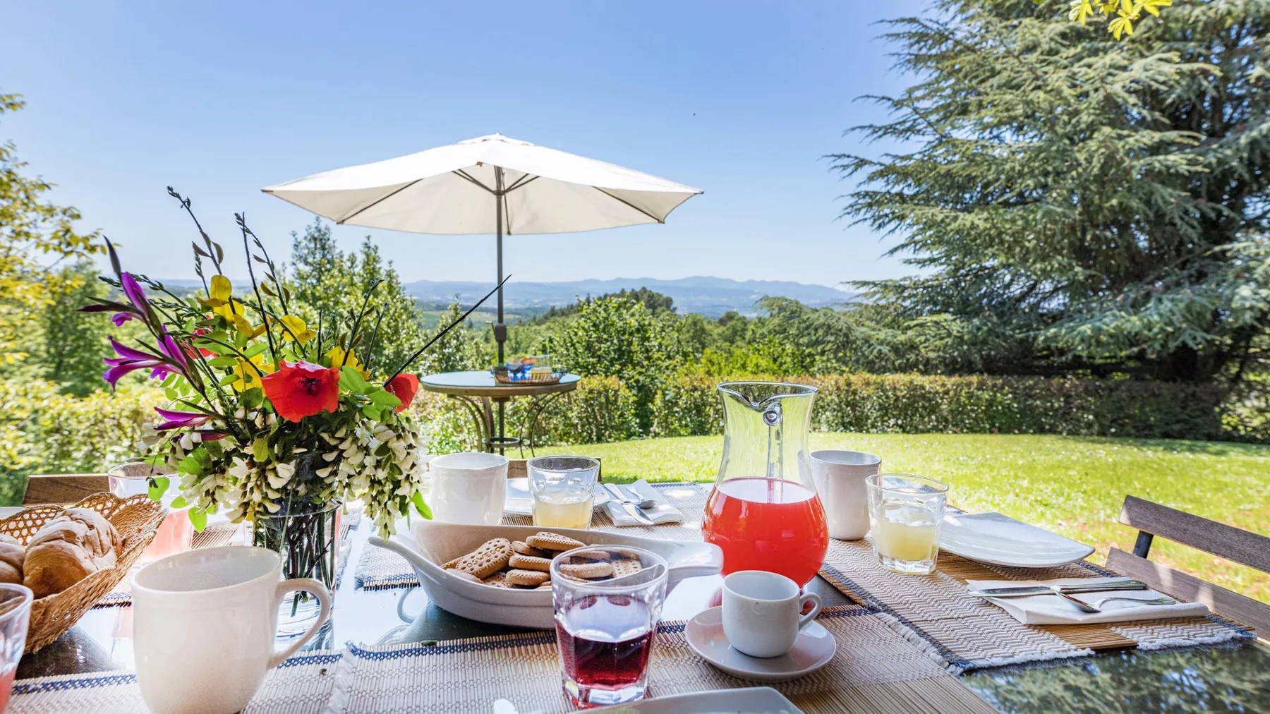 Una mesa al aire libre está preparada con bebidas y comida bajo una sombrilla blanca, frente a un paisaje verde.