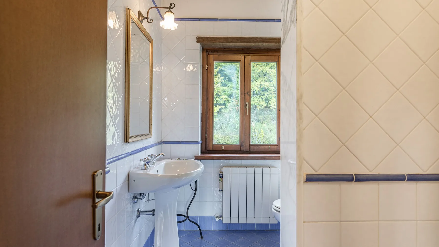 Un baño alicatado con lavabo de pedestal y una ventana con vistas a la vegetación.