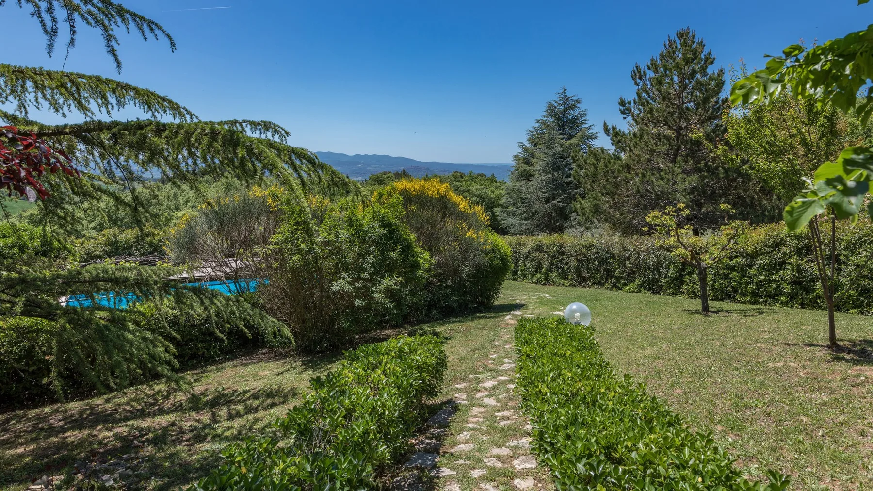 Un sendero de jardín atraviesa la vegetación, con árboles y colinas lejanas bajo un cielo despejado.