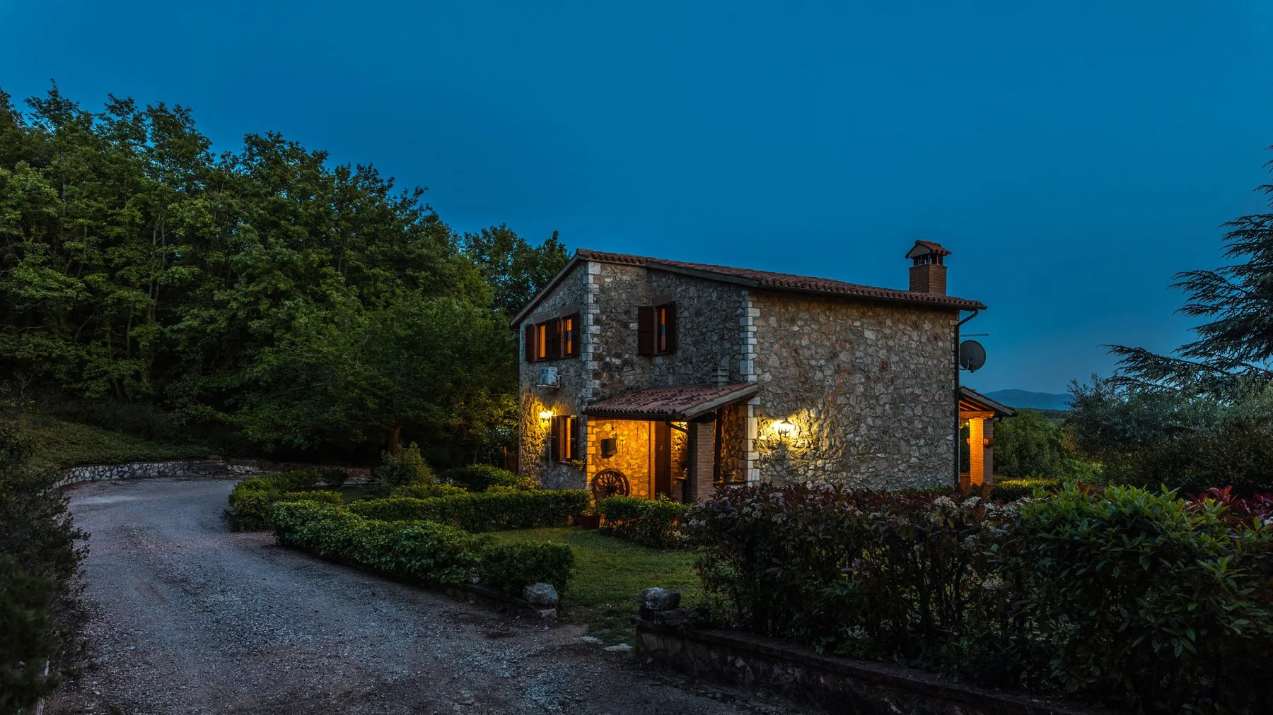 Una casa de piedra brilla con luces cálidas junto a un camino de grava al anochecer.