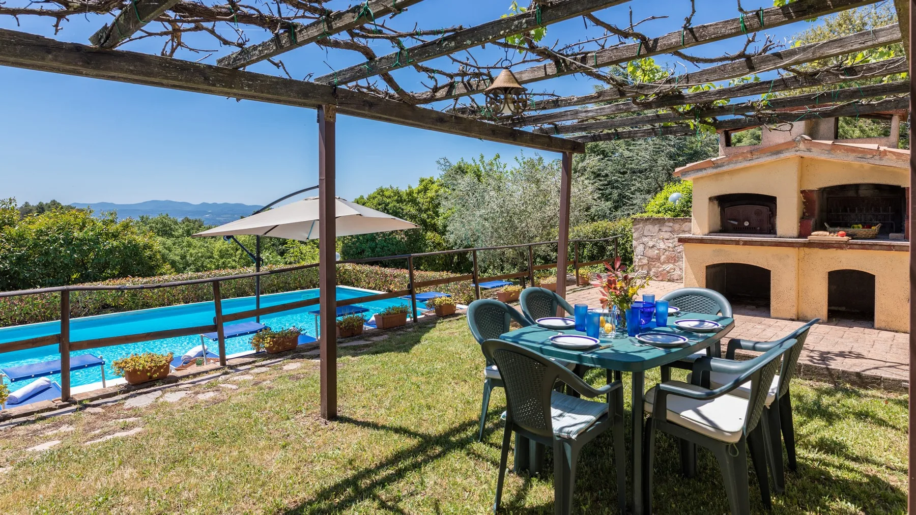 Una zona de comedor al aire libre bajo una pérgola da a una piscina y un horno exterior.