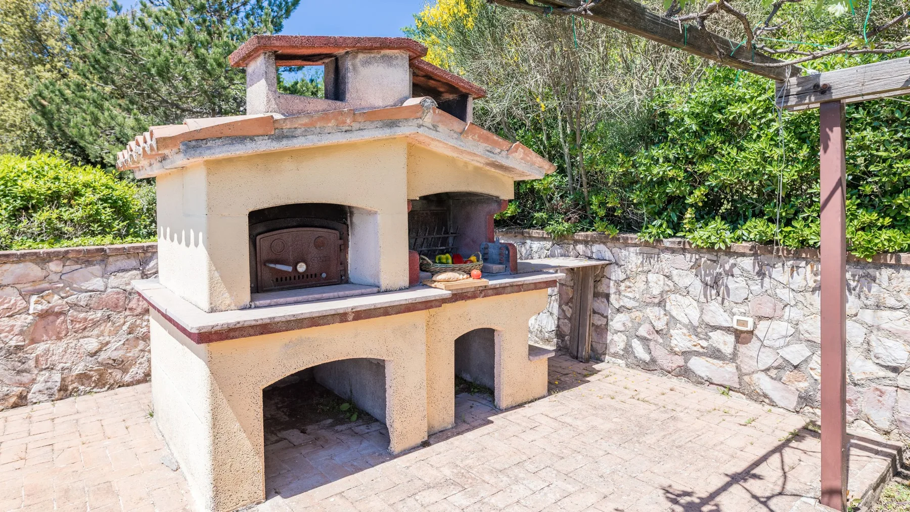 Una barbacoa de obra al aire libre está en un patio pavimentado junto a un muro de piedra.