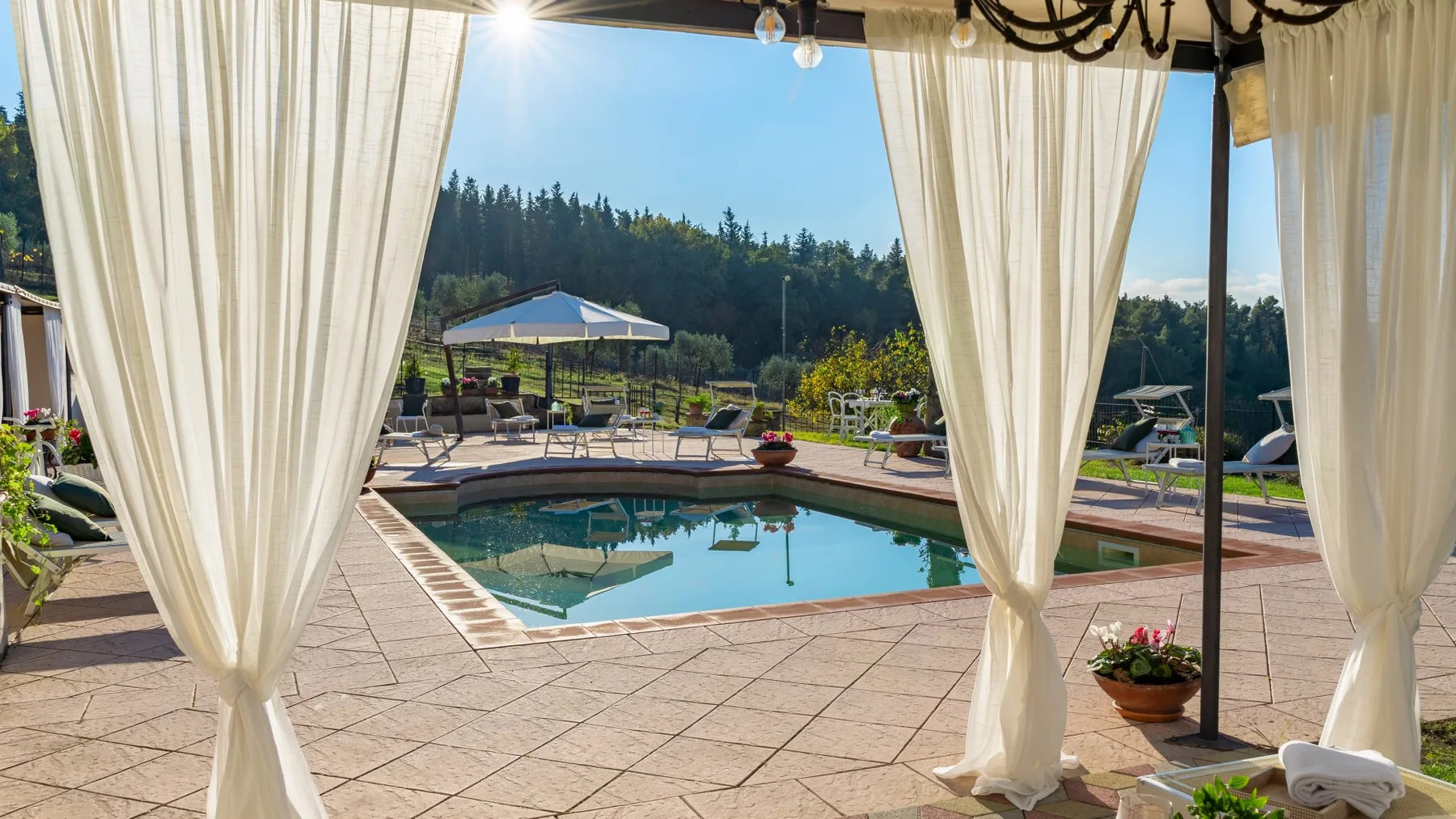 Un patio con piscina de agua turquesa, asientos al aire libre y cortinas que enmarcan la vista.
