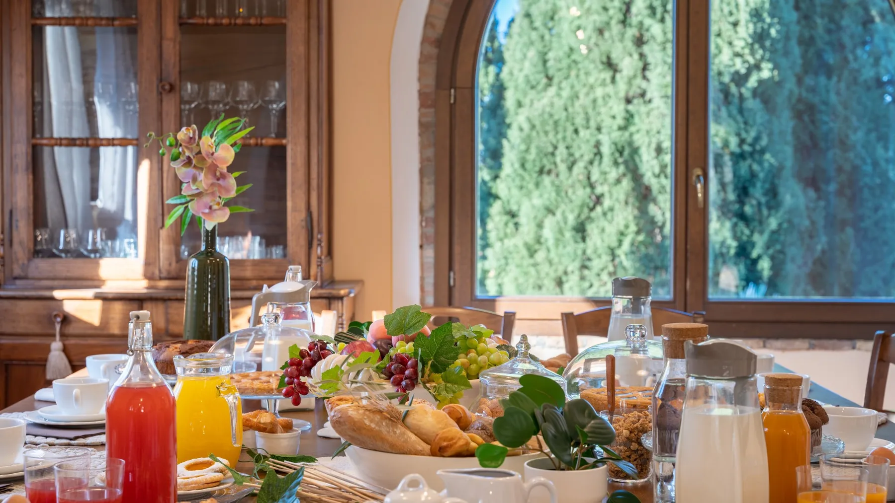 Una mesa de comedor está dispuesta con pan, fruta, bebidas y flores junto a una ventana arqueada.