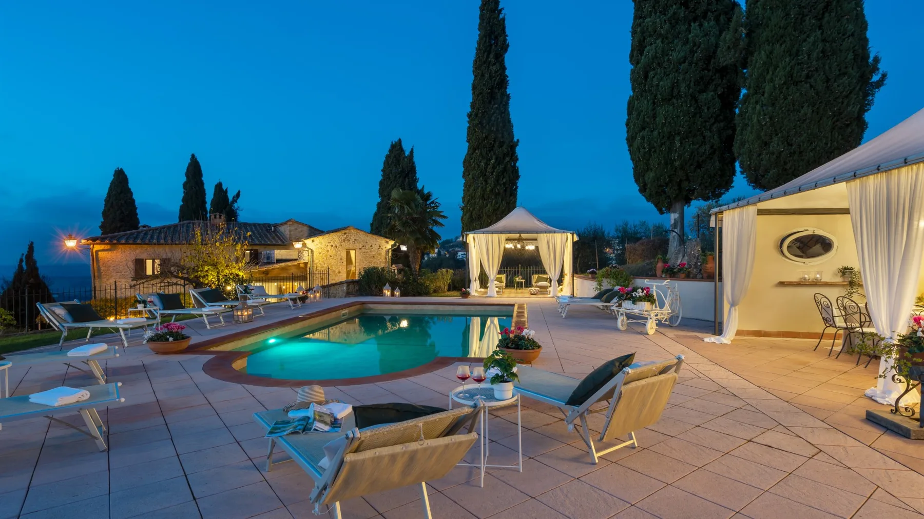 Una piscina al aire libre y asientos en el patio están iluminados bajo el cielo azul del anochecer.