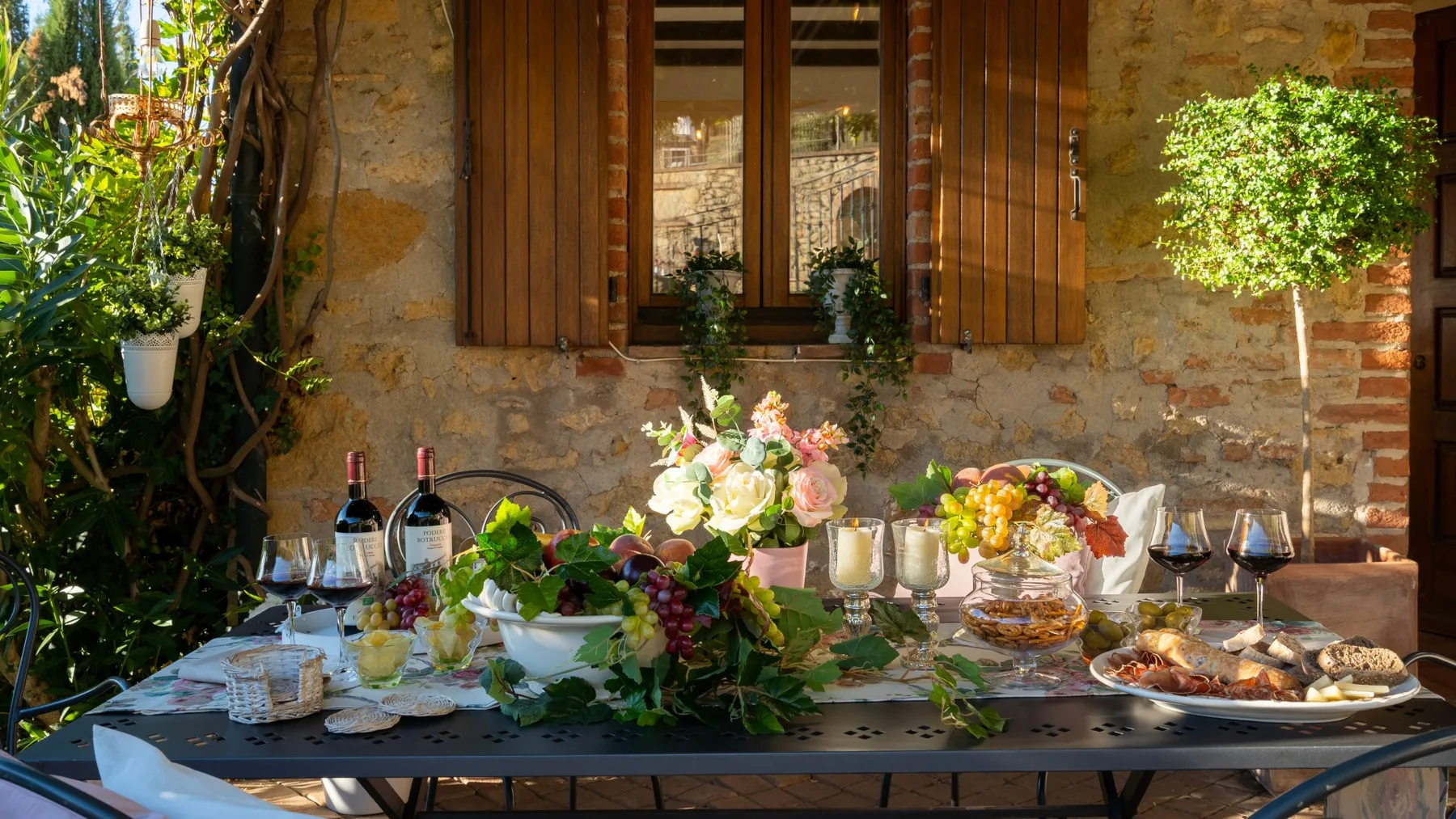 Una mesa de comedor al aire libre está preparada con comida, vino, flores y plantas.