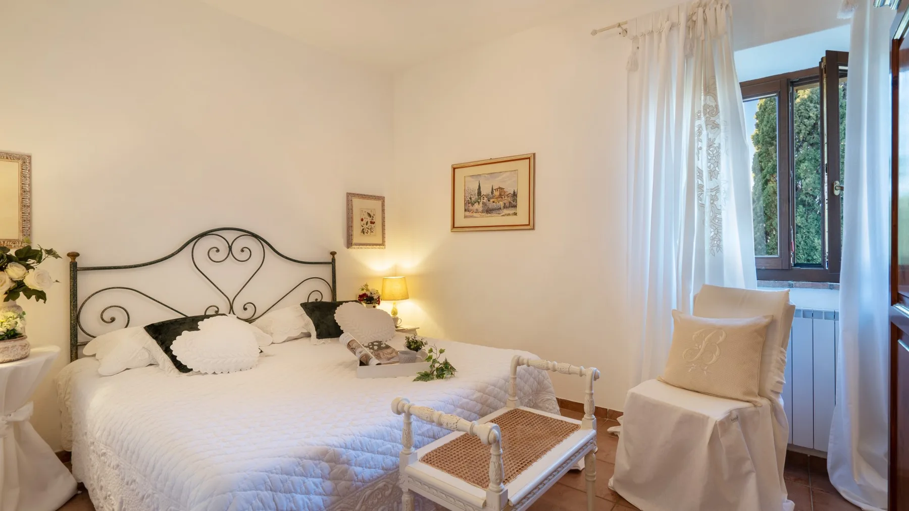 Un dormitorio con una cama blanca, una lámpara de noche y una ventana con cortinas translúcidas.
