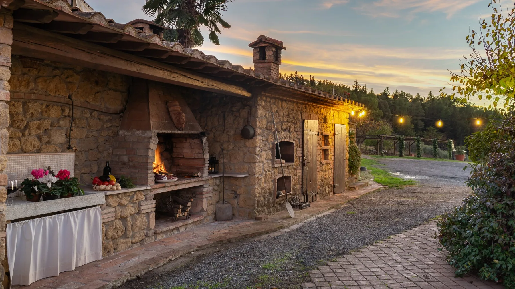 Una zona de cocina exterior de piedra da a un patio pavimentado al atardecer.