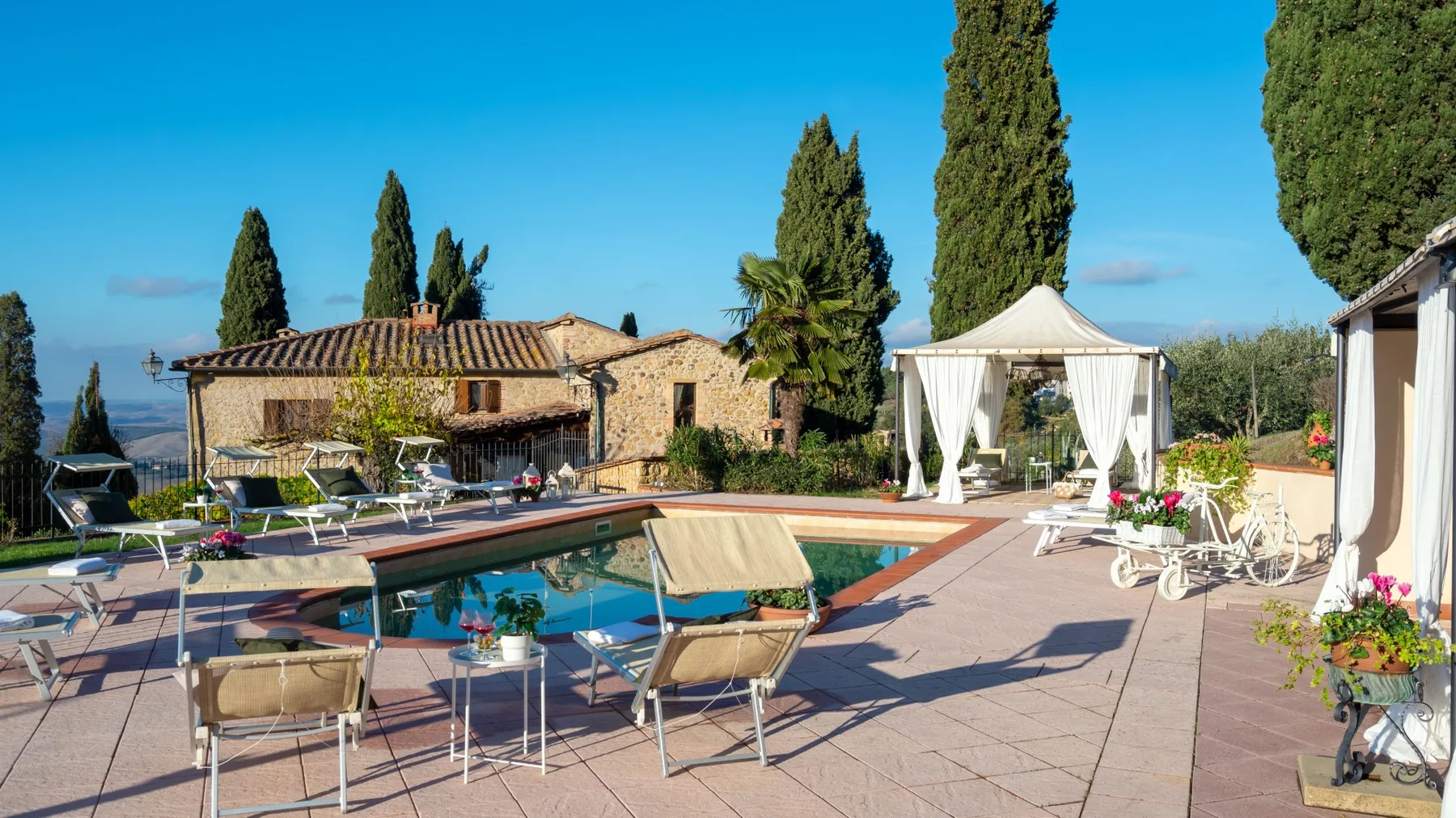 Una piscina al aire libre junto a un edificio de piedra, con asientos y una carpa blanca.