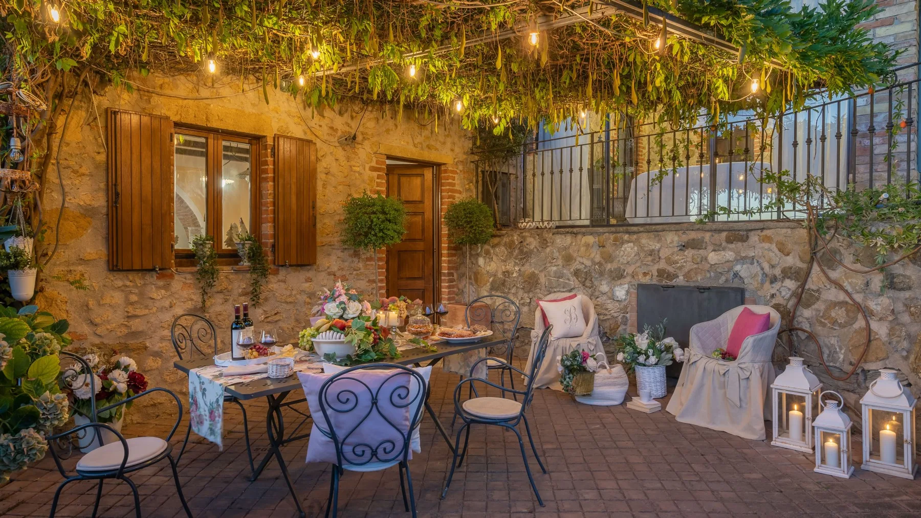 Un patio de piedra con zona de comedor, asientos, plantas y cálidas luces colgantes.