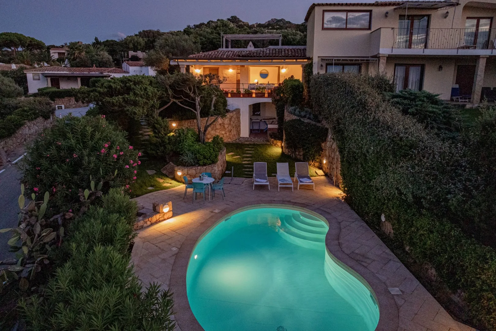 A lit outdoor pool sits among landscaped gardens beside a residence at dusk.