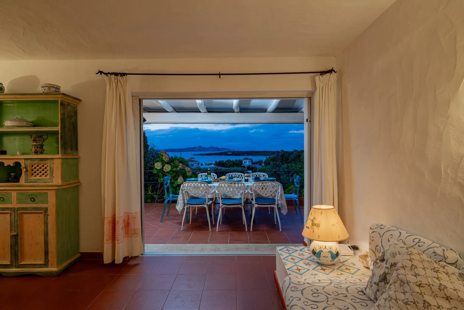 A living room opens onto a terrace with a dining table and a distant sea view.