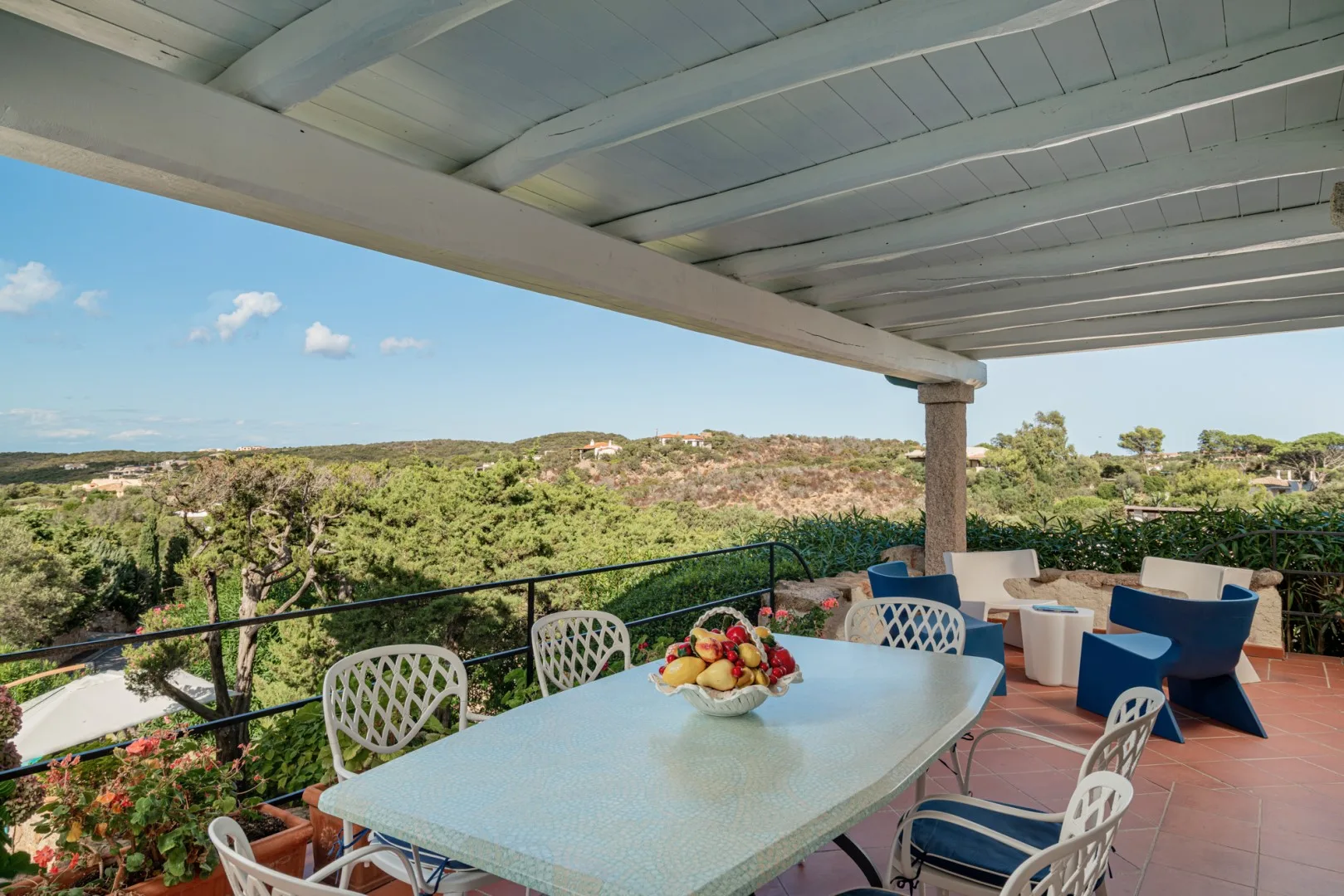 A covered terrace has a dining table and chairs overlooking a green hillside.