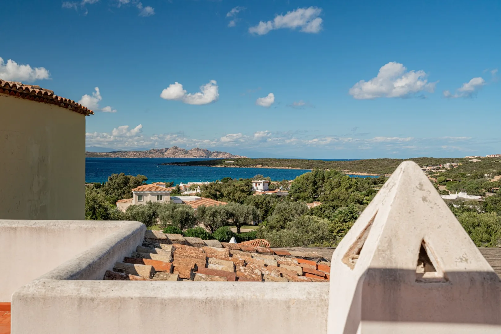 A rooftop terrace overlooks greenery, nearby homes, and blue water beneath a clear sky.