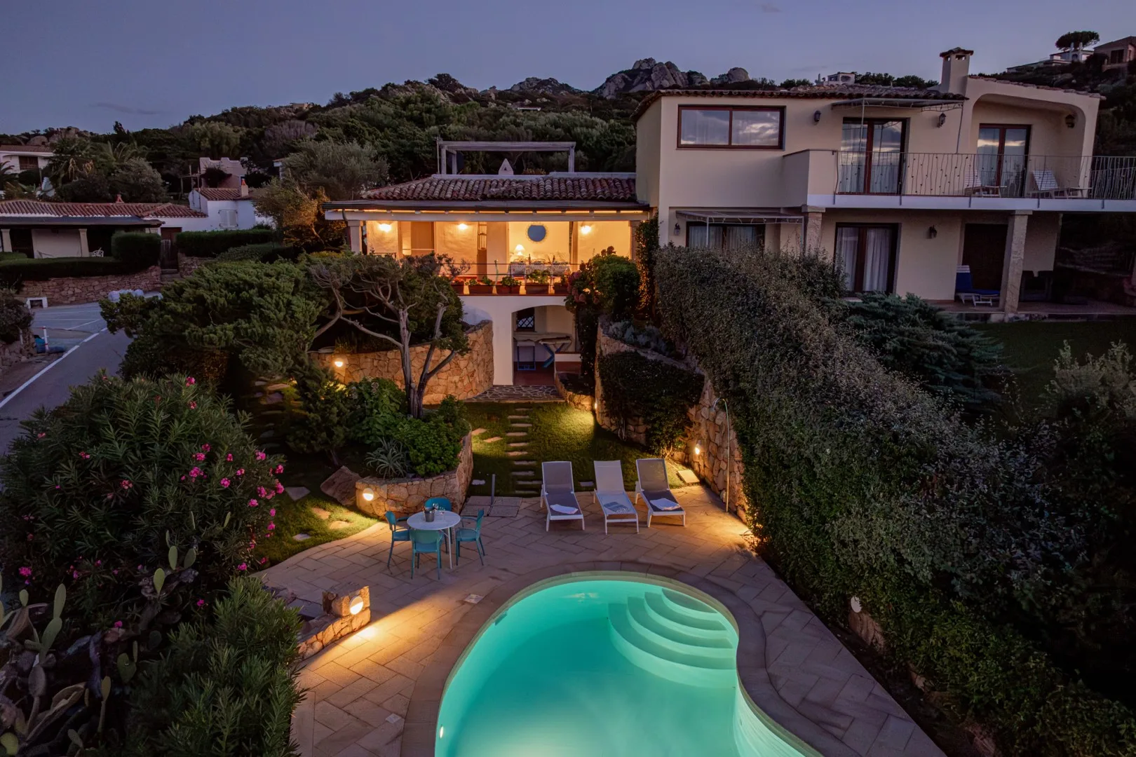 A villa and landscaped garden overlook a lit swimming pool at dusk.