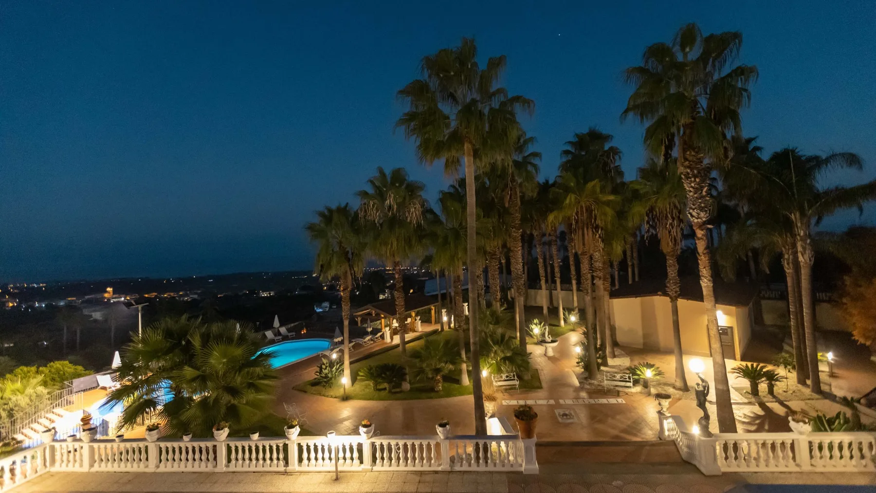 Una zona exterior ajardinada de noche, con palmeras y senderos iluminados.