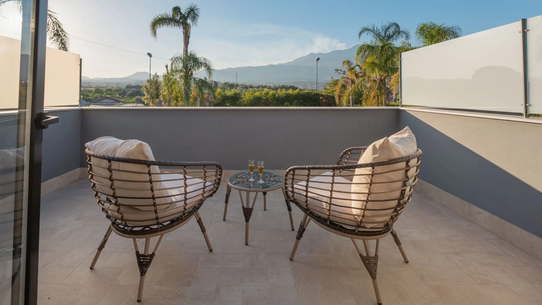 Una terraza en la azotea con dos asientos acolchados y una mesa pequeña, frente a palmeras y colinas.