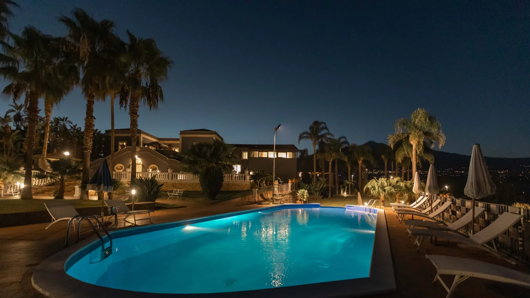 Una piscina exterior iluminada está rodeada de tumbonas y palmeras por la noche.