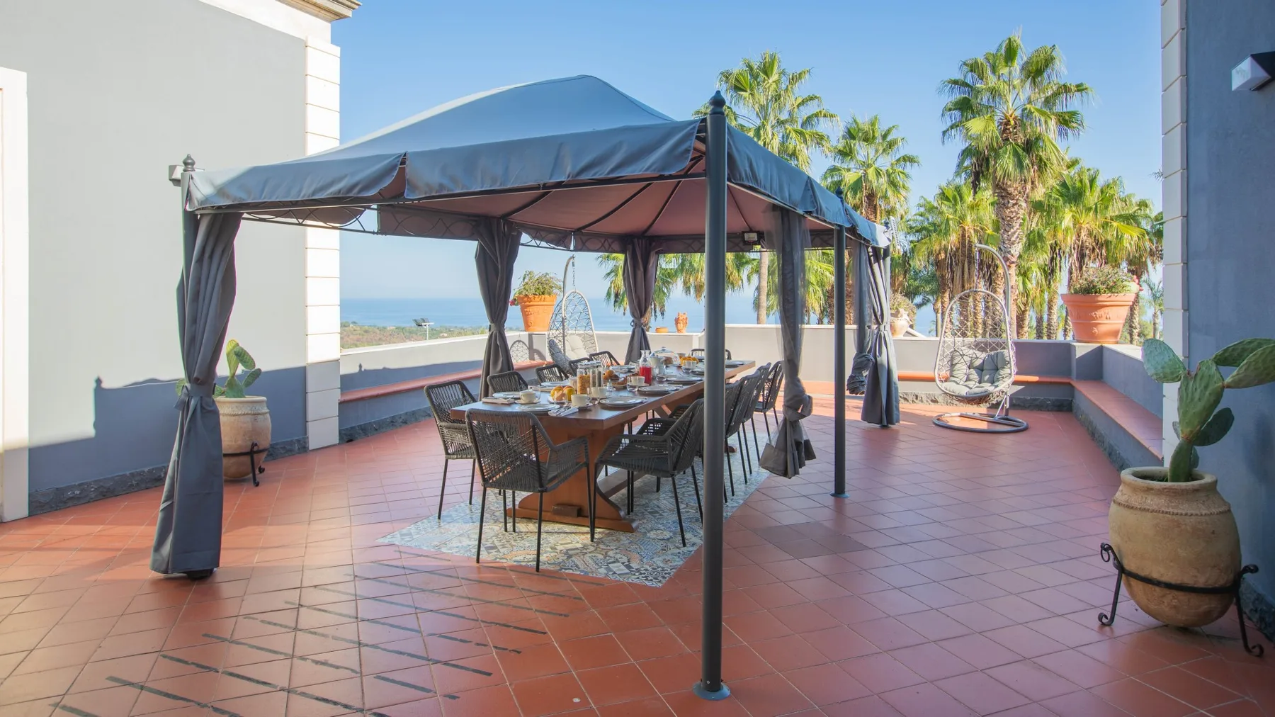 Una terraza exterior de baldosas tiene una mesa de comedor bajo una cubierta de tela, con palmeras cerca.