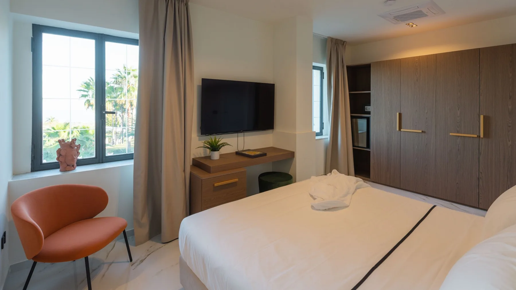 Un dormitorio con una cama blanca, un televisor y una ventana con vistas a la vegetación.