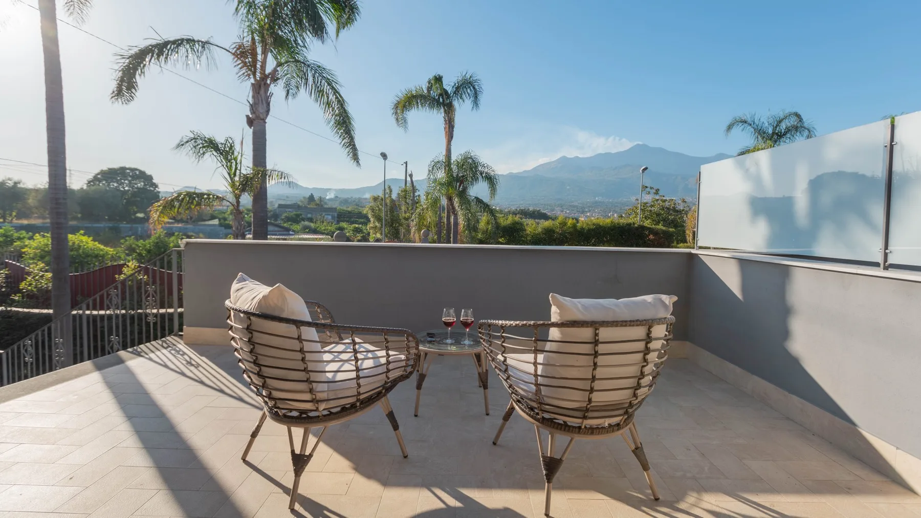 Una terraza soleada tiene asientos y una mesa pequeña, con palmeras y montañas al fondo.