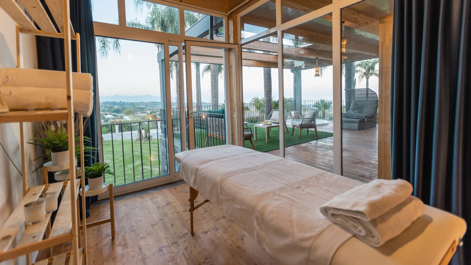 Una camilla de masaje está junto a ventanales con vistas a un patio ajardinado.