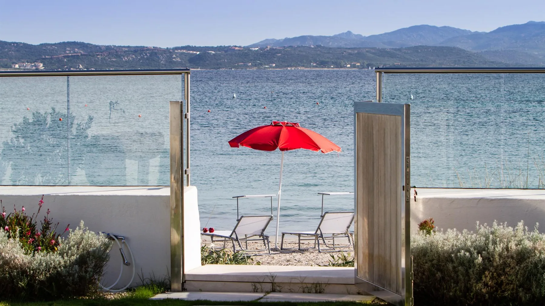 Un patio junto a la playa se abre al agua, con una sombrilla roja sobre asientos.