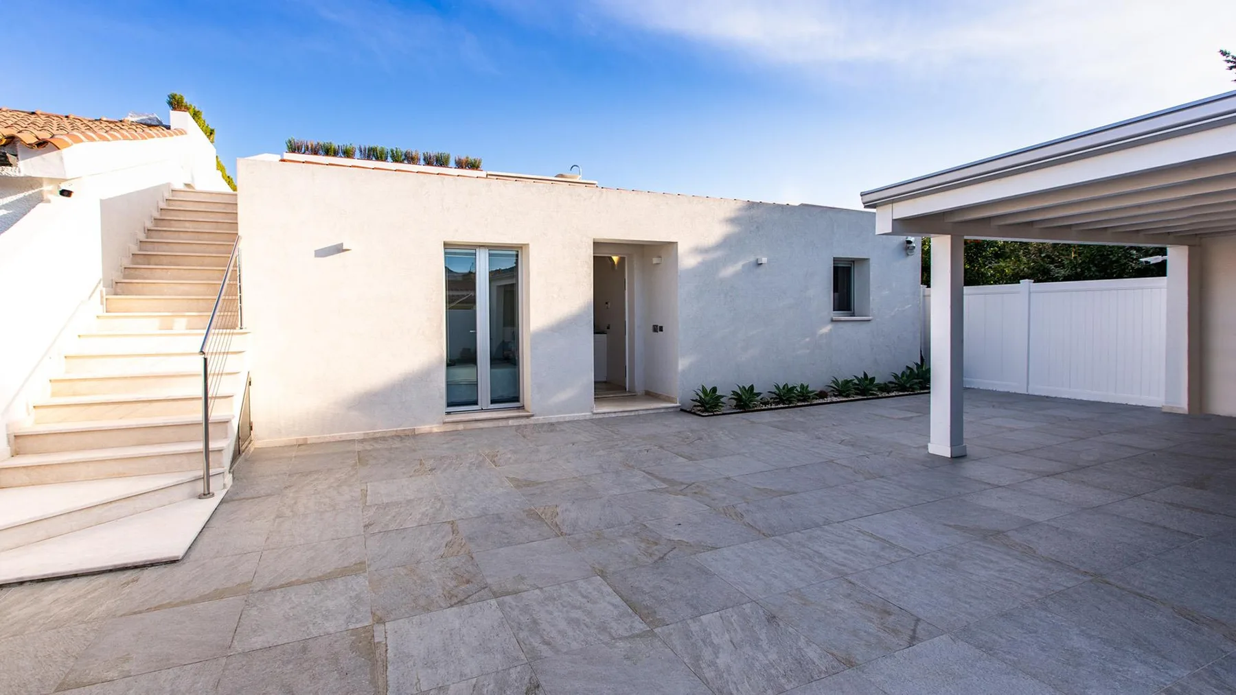 Un patio pavimentado junto a un edificio blanco, con una escalera exterior y una zona de estacionamiento cubierta.