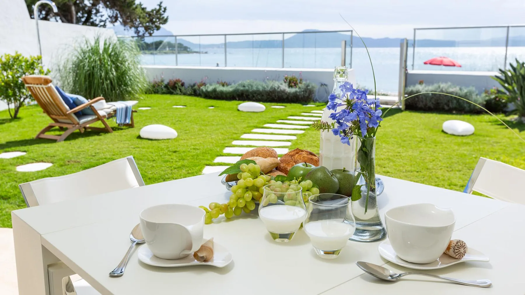 Una mesa de jardín está puesta con tazas, fruta y flores junto a un césped verde.