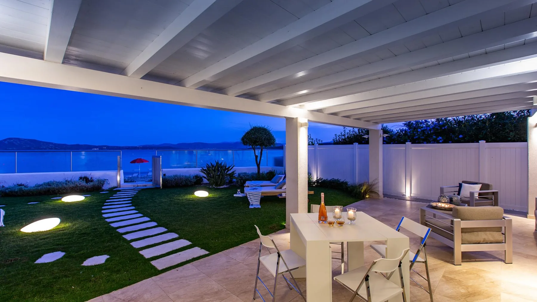 Una zona de comedor en un patio cubierto se abre a un césped junto a la costa.