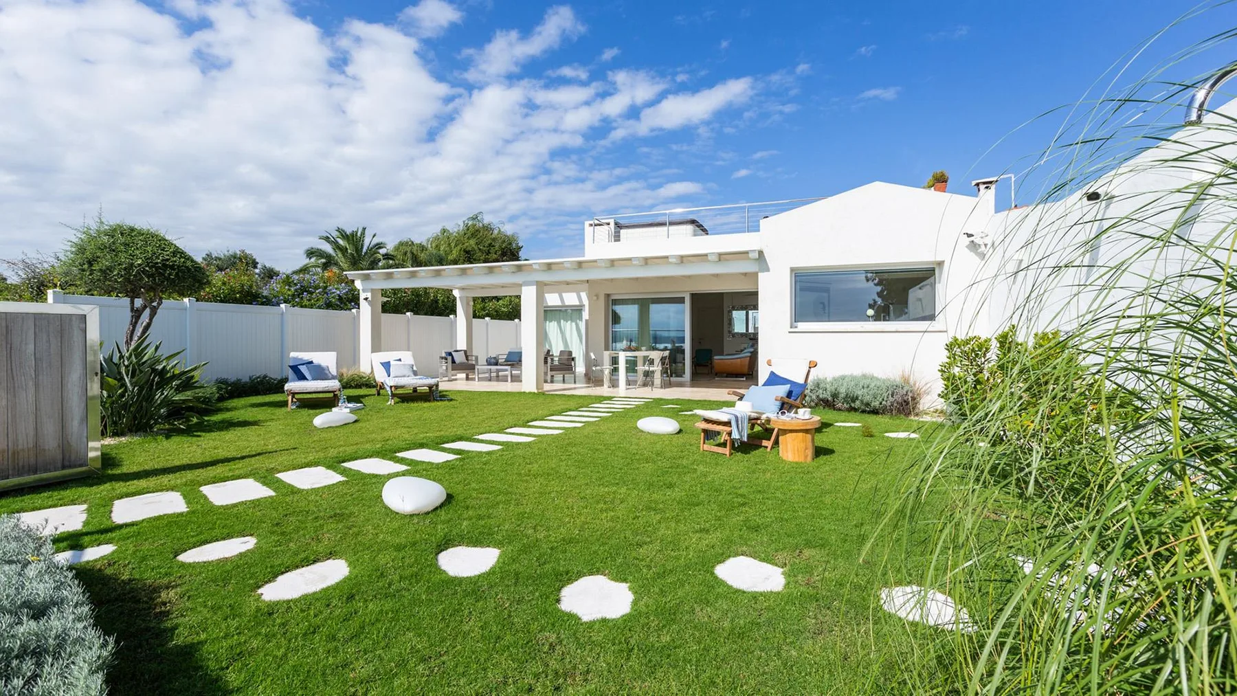 Una casa blanca moderna da a un césped verde con piedras de paso y asientos en el patio.