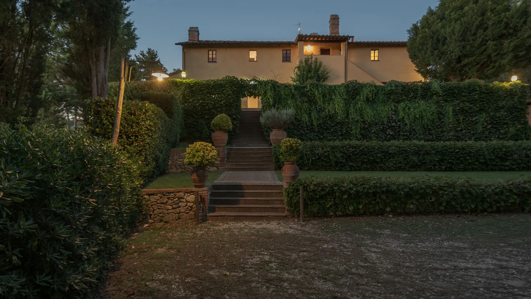 Un sendero de jardín y unos escalones atraviesan los setos hacia una casa iluminada al anochecer.