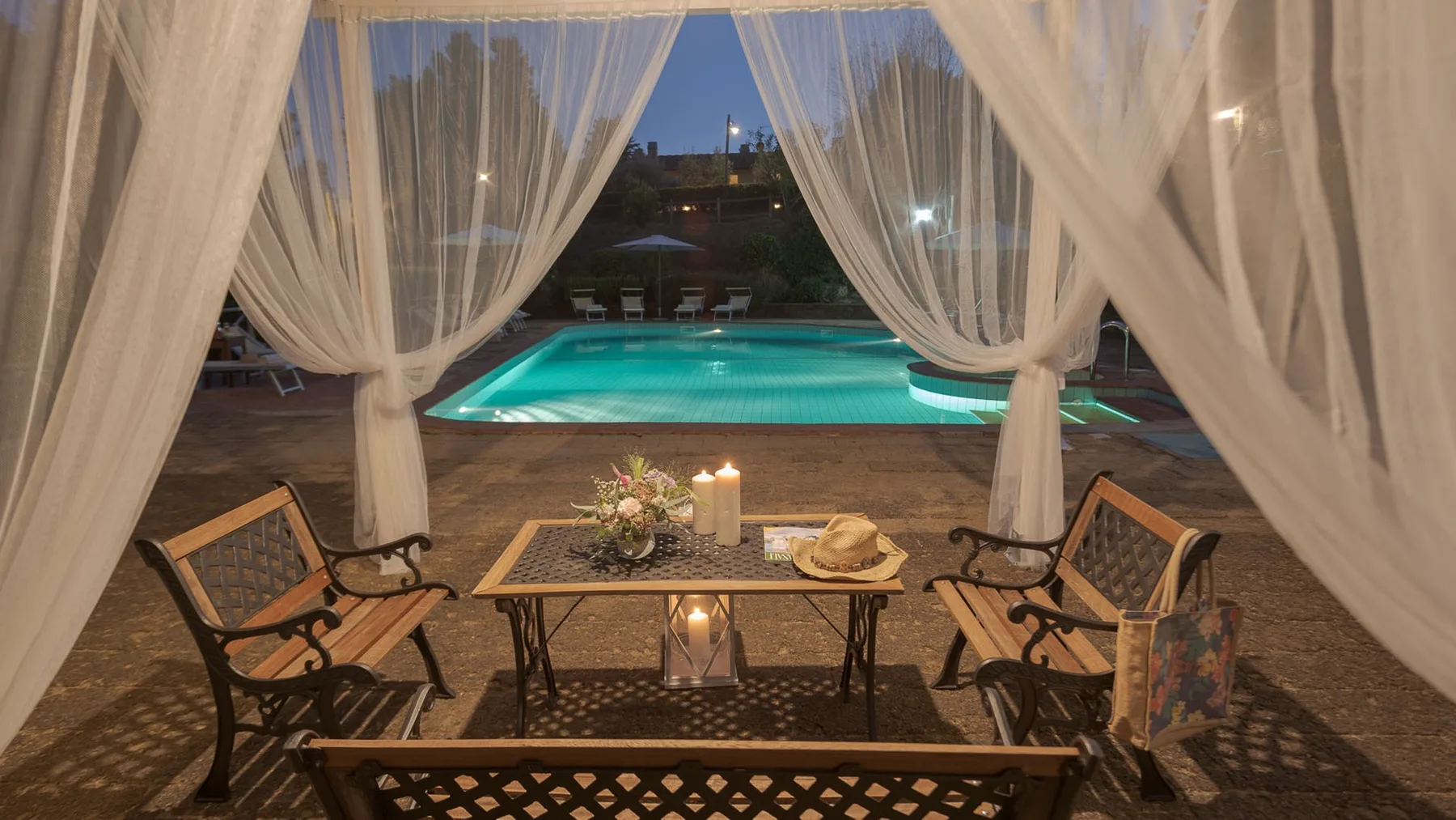 Una zona de estar al aire libre con cortinas da a una piscina iluminada al anochecer.