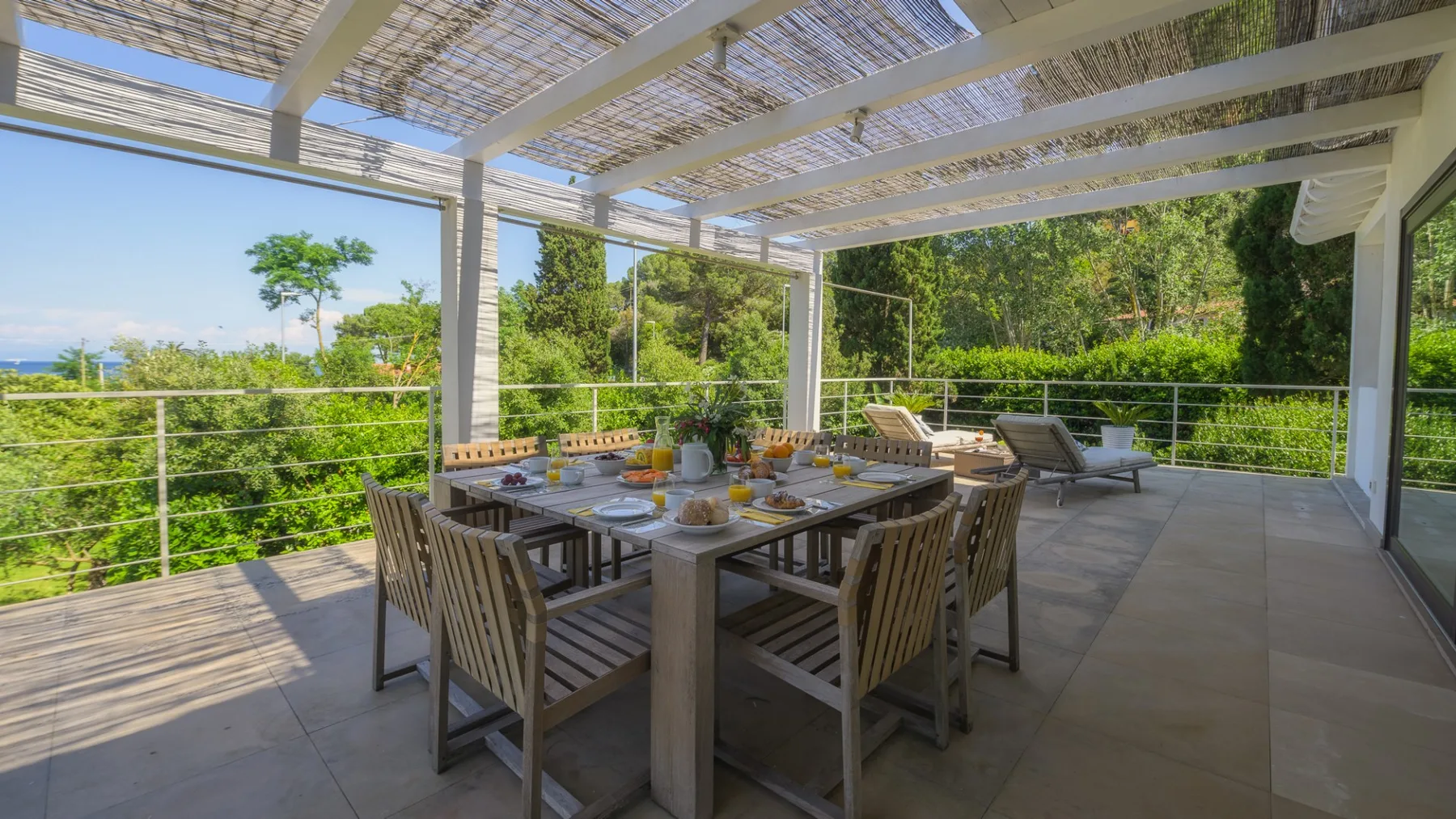 An outdoor dining area sits beneath a shaded pergola, surrounded by greenery.
