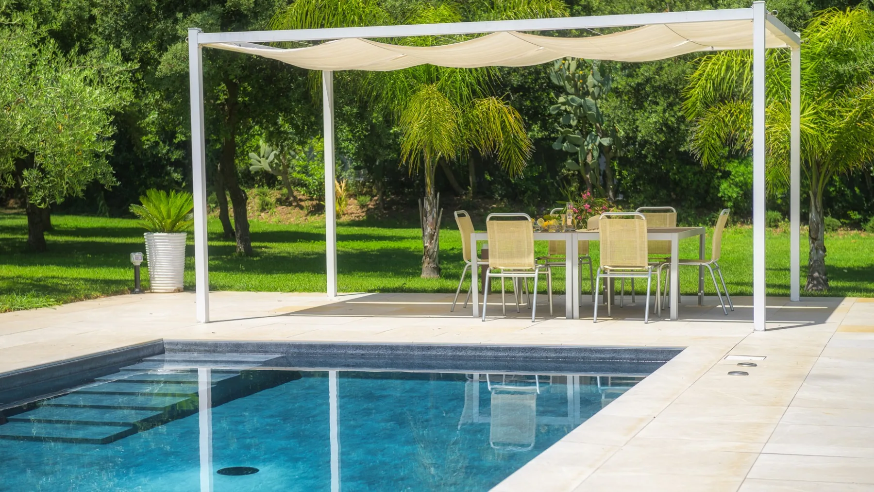 An outdoor pool sits beside a shaded patio dining area surrounded by greenery.