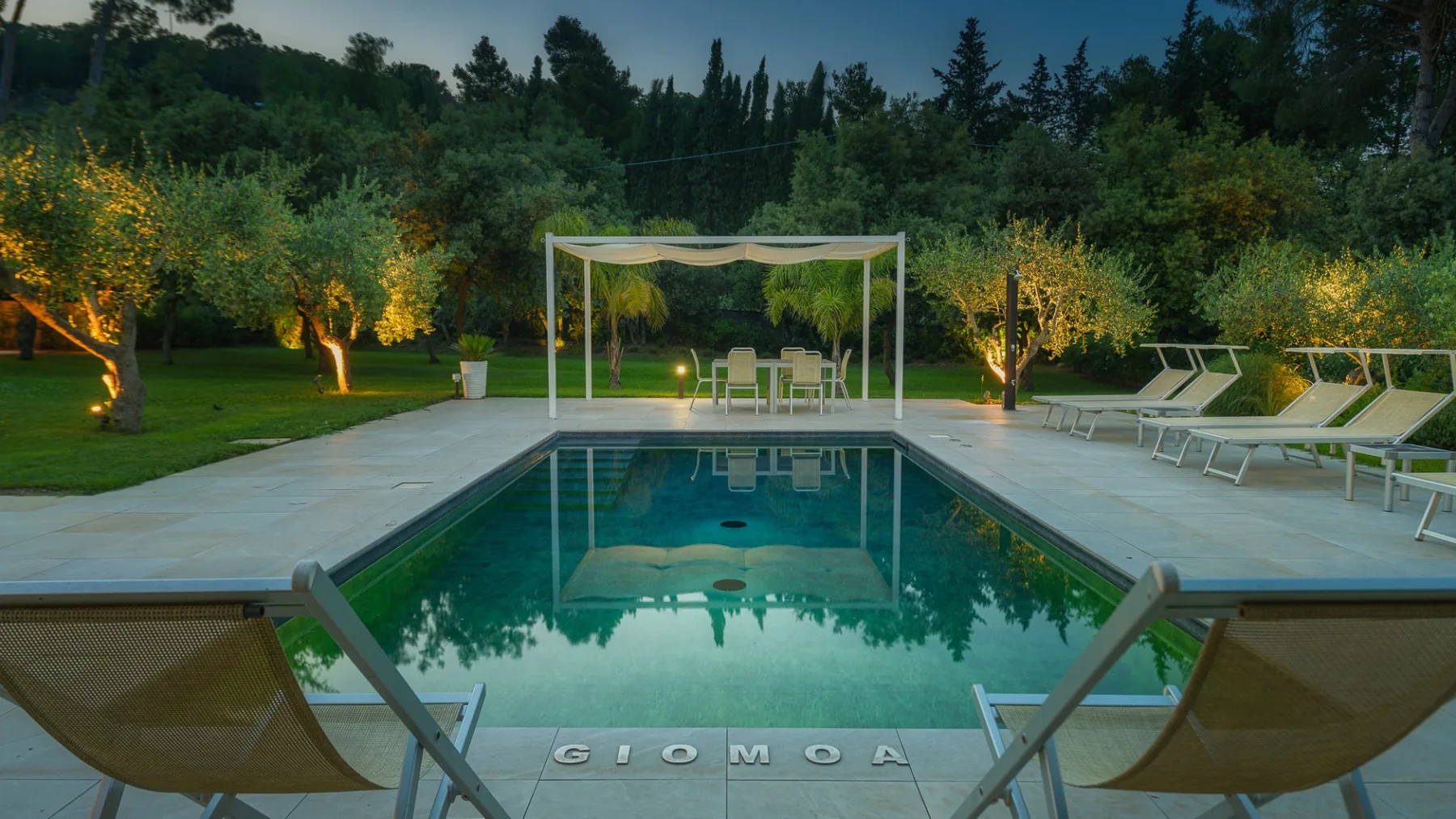 An outdoor swimming pool faces a shaded seating area among landscaped trees.