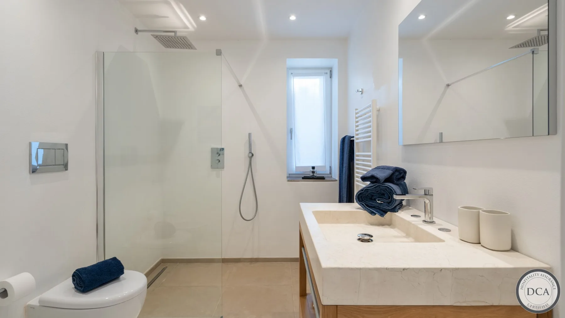 A bright bathroom with a glass shower, vanity, and a small window.