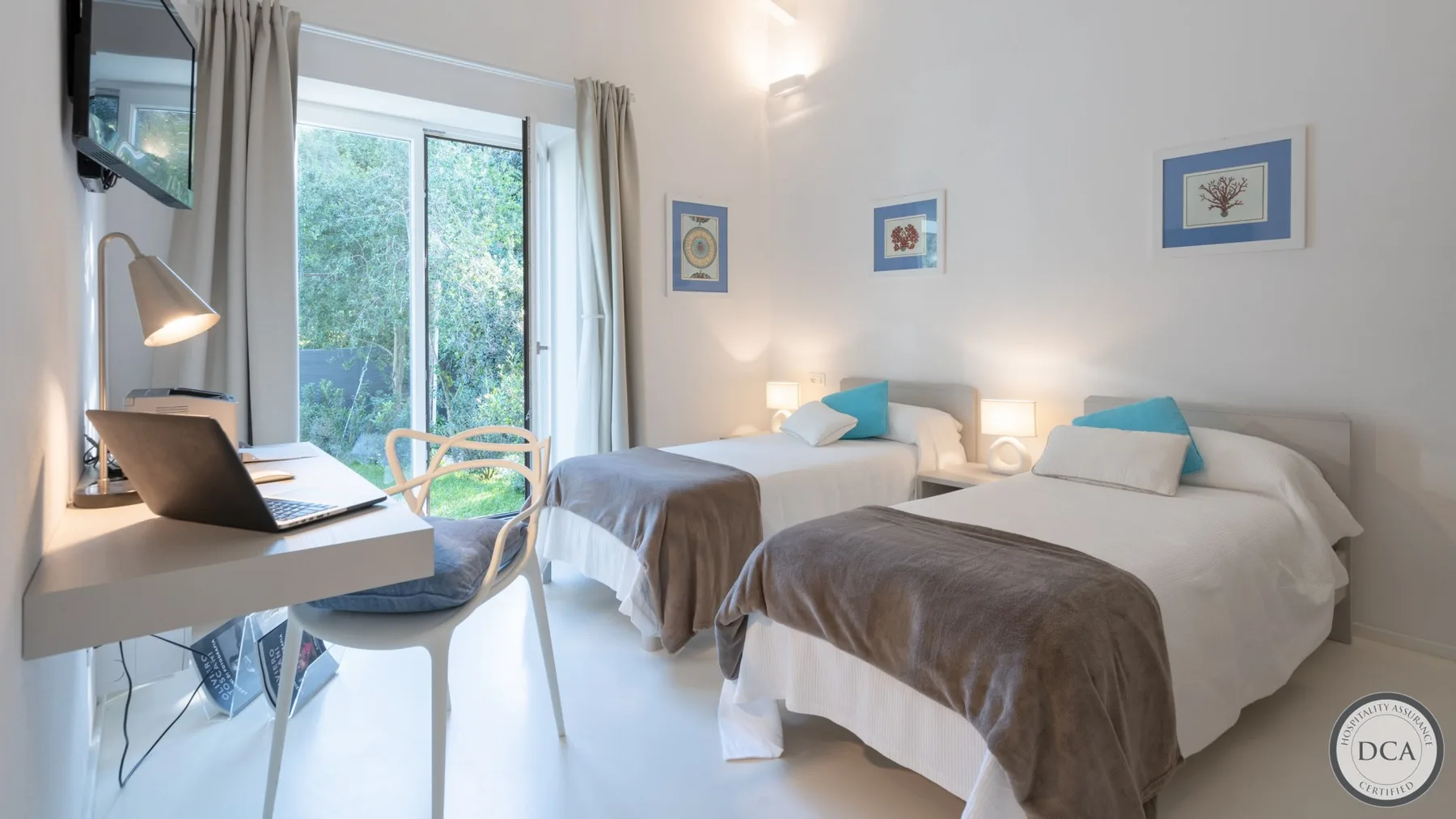 A bright bedroom with two beds and a desk beside a glass door.