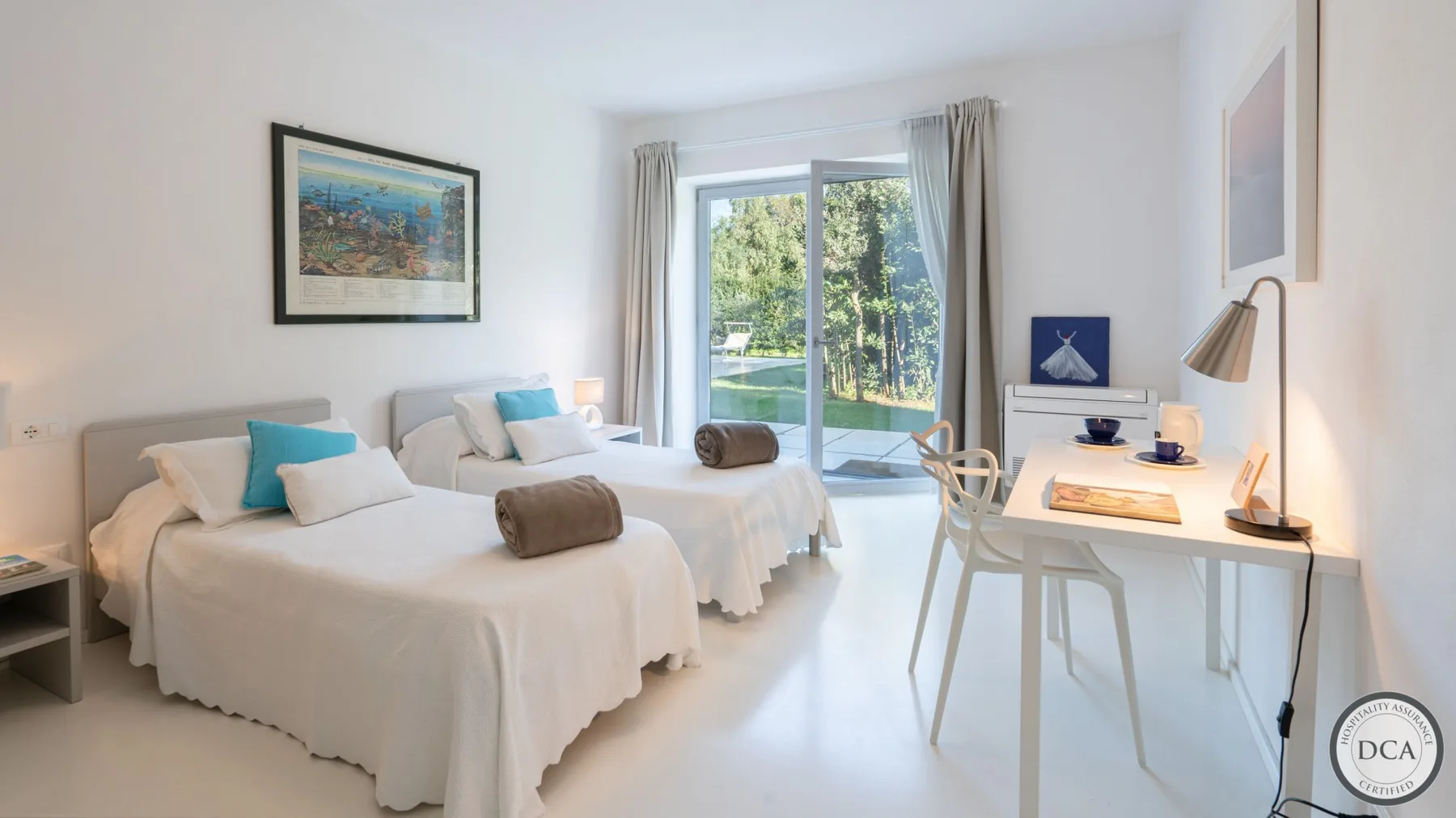A bright bedroom with two beds and a desk beside a glass door.