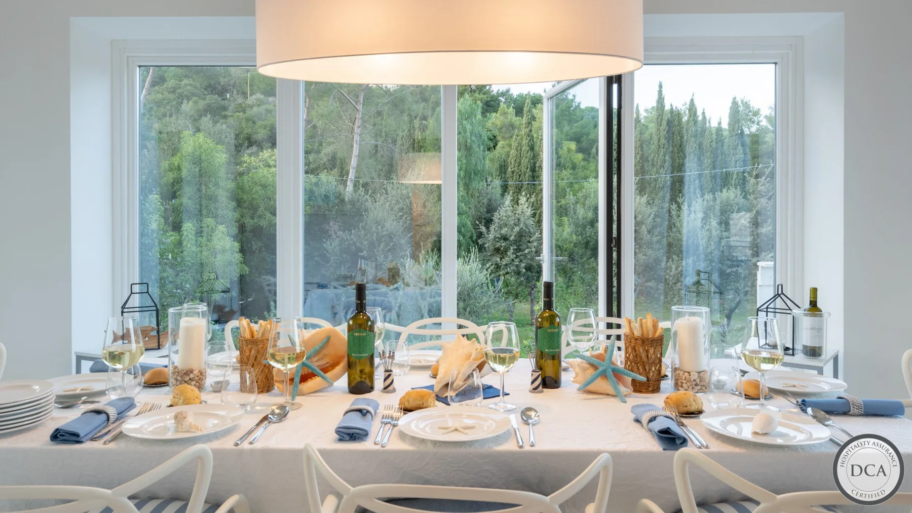 A dining table is set beside large windows overlooking a wooded landscape.