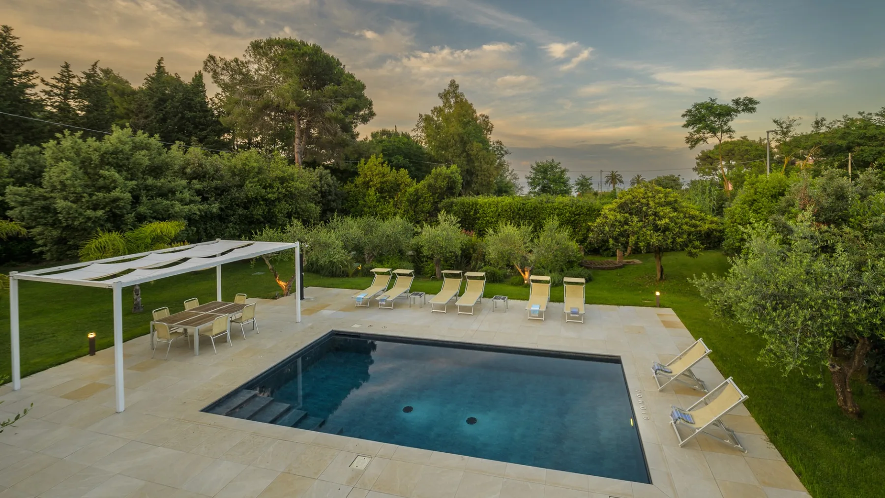 An outdoor swimming pool sits beside a paved patio with seating and loungers.
