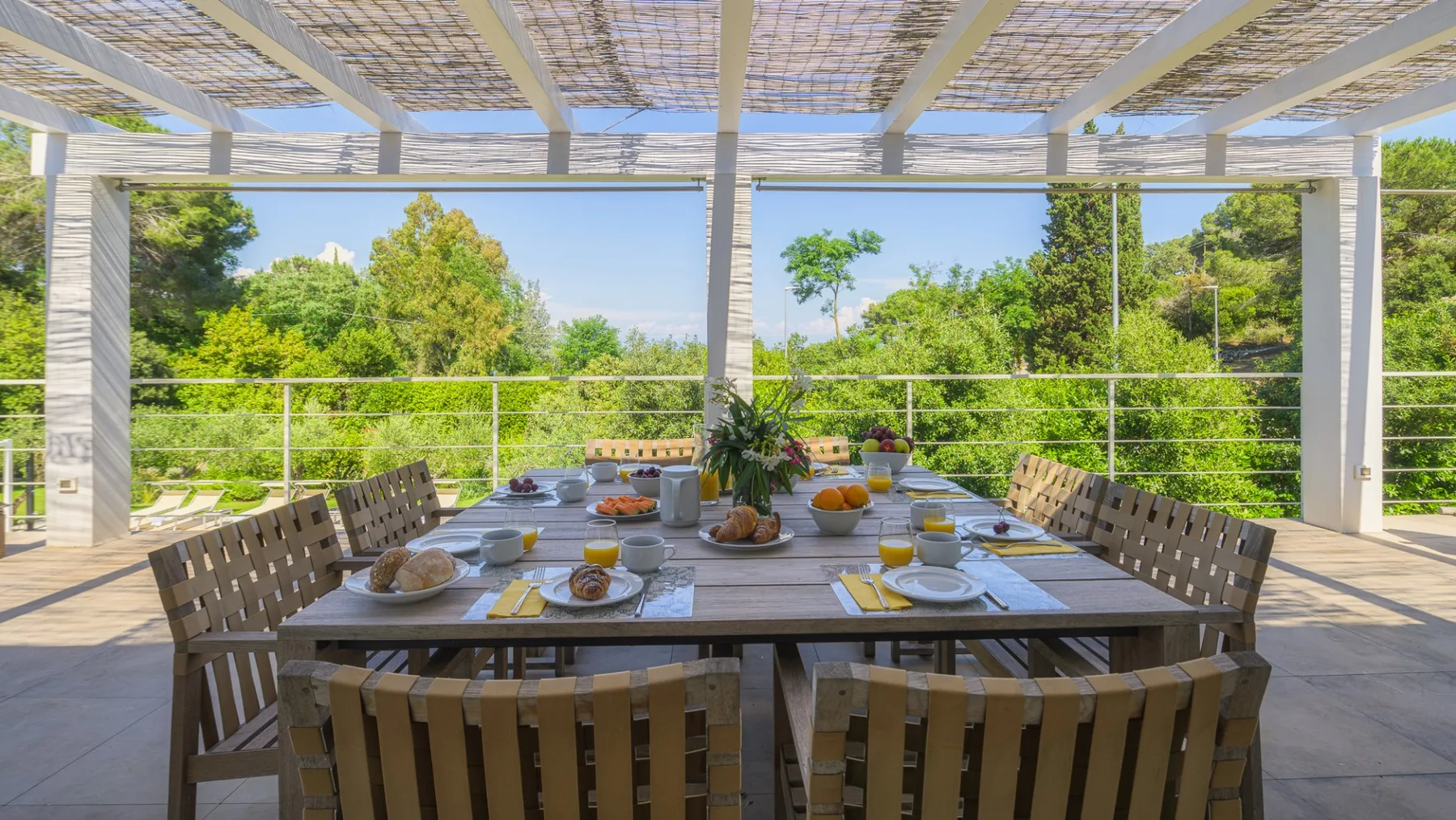 An outdoor dining area sits beneath a pergola, with a long table and surrounding greenery.