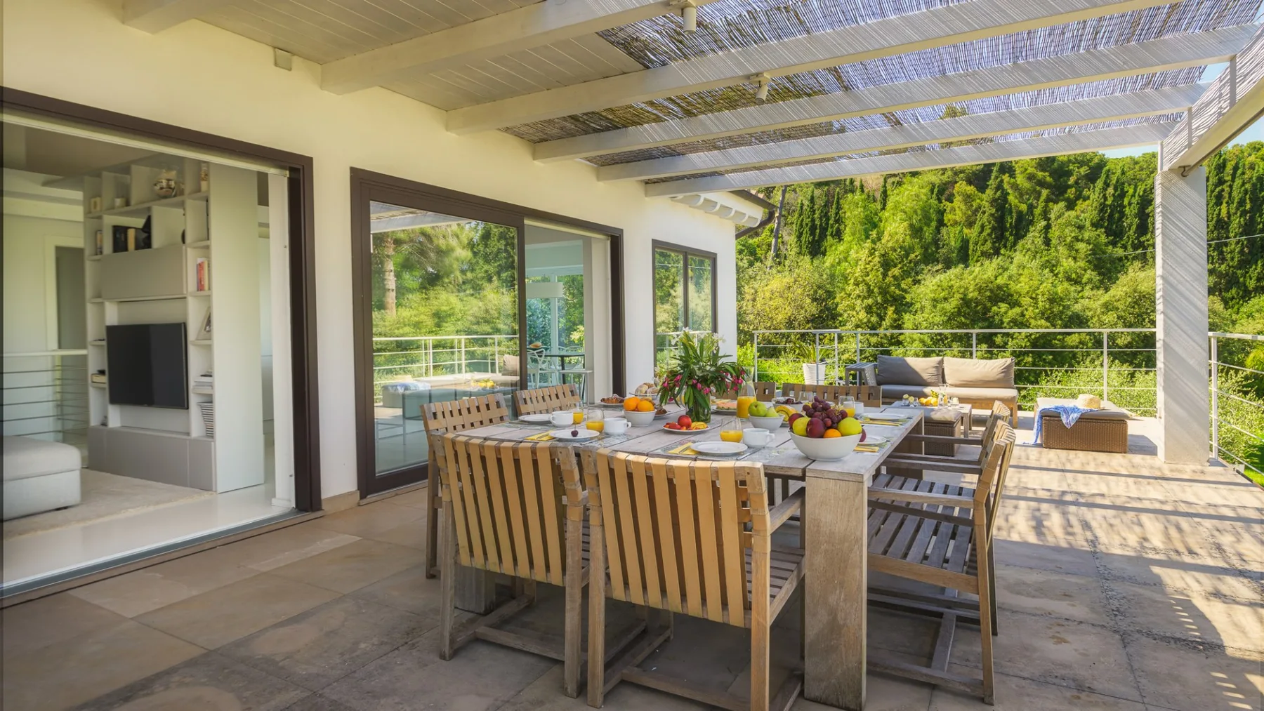 An outdoor dining area with a long table and greenery beyond the terrace.