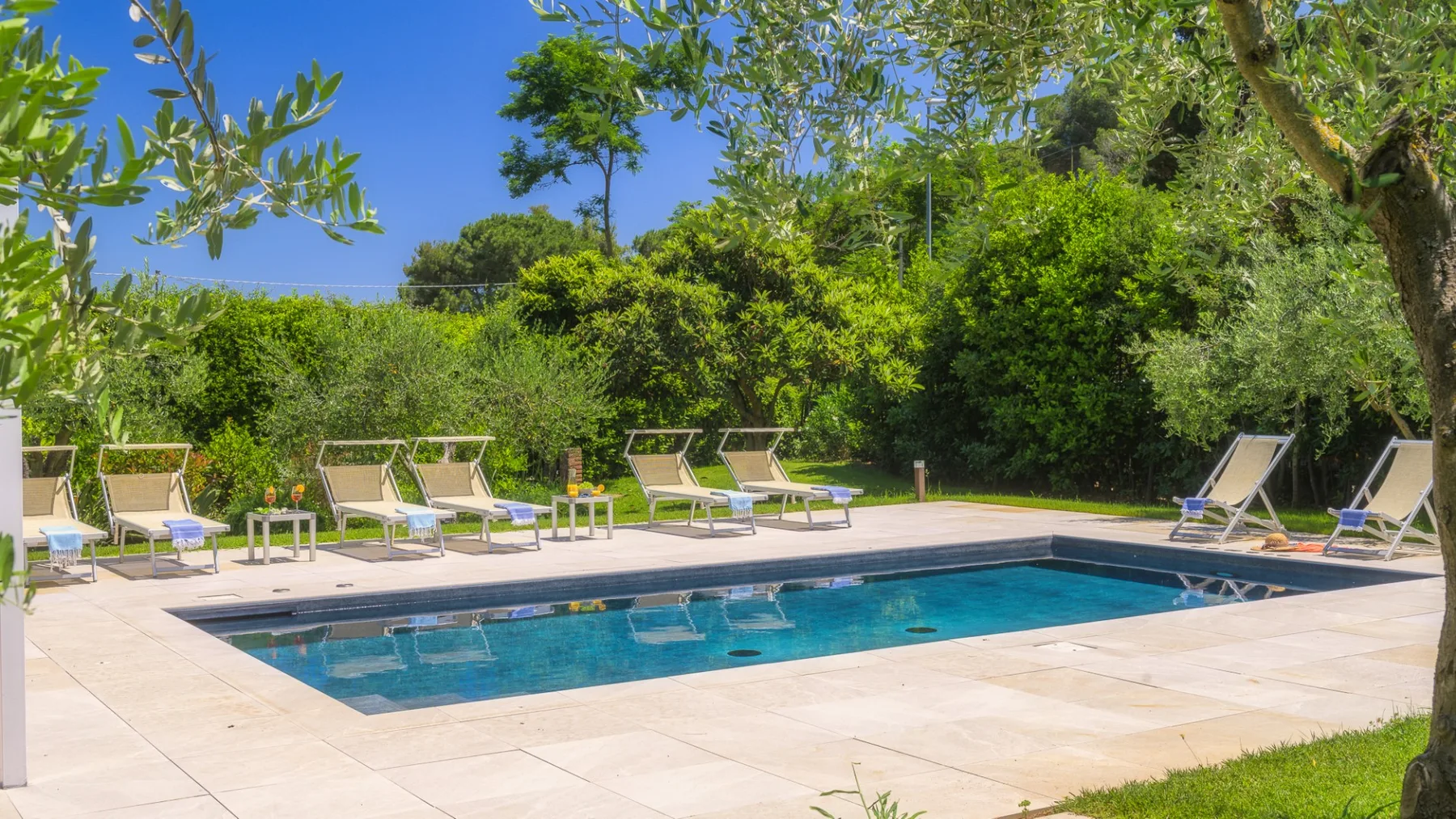 An outdoor swimming pool sits beside a paved deck with lounge seating and greenery.